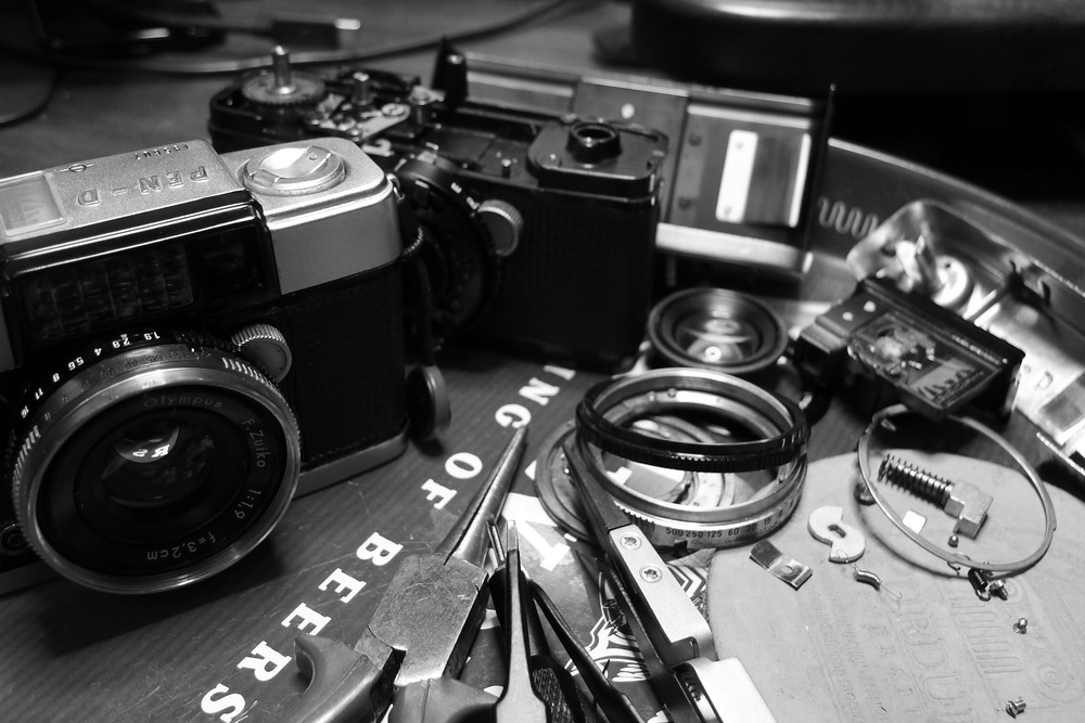 two olympus pen d cameras, one disassembled, with parts and tools scattered around a tray