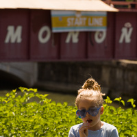 A girl in blue hear-shaped glasses eating a lollipop in front of a pale red bridge that says 