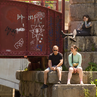 A few people sit on the abutments of a red former railway bridge.