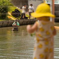 Officials and volunteers wait patiently at the finish line, one standing in the canal in waders. In the foreground, a small child dressed in a bright yellow hat and duck-themed clothing stands at the railing watching.